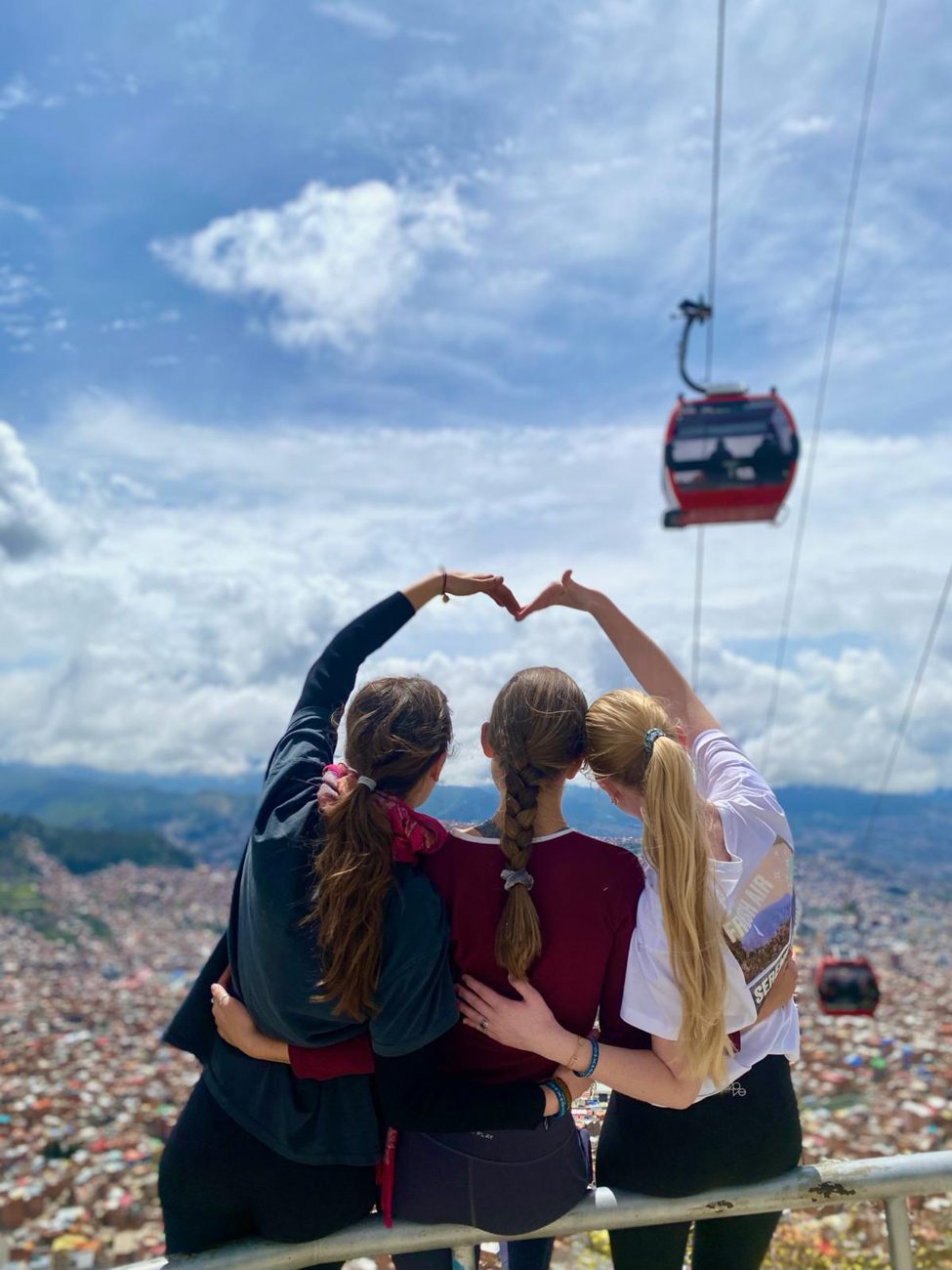 Tres jóvenes forman un corazón con las manos, al fondo La Paz con el teleférico
